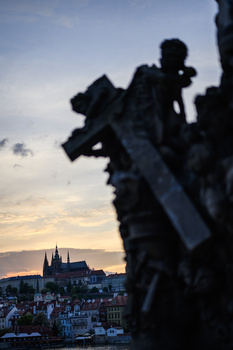 Silhouetted statue in foreground overlooking a European city at golden hour with a cathedral visible across the river.