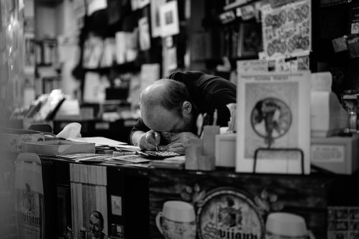 Person with dark hair leaning over a desk covered with papers and objects in a black and white photograph.