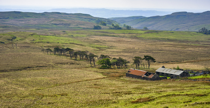 Rural hillside valley with scattered farm buildings, tree-lined stream, and rolling mountains under overcast sky.