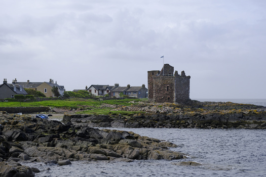 Stone castle tower on rocky coastline with village houses and overcast sky.