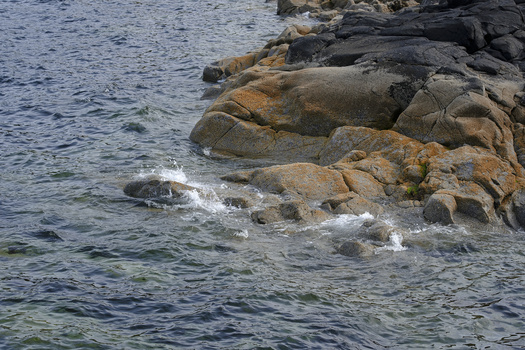 Waves crash against a rocky shoreline scattered with large boulders under bright sunlight.