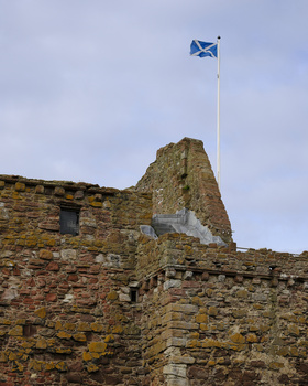 Scottish flag flying from a pole atop weathered stone castle ruins under overcast sky.