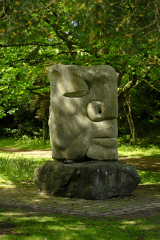 Stone sculpture of an abstract human head mounted on a pedestal in a park setting.
