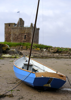 Blue and white sailboat beached on sand with stone tower ruins on hillside in background.