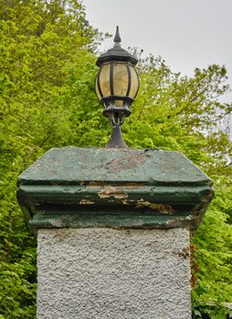 Vintage-style lantern mounted atop a weathered stone post surrounded by dense green foliage.