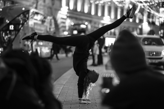 Silhouetted person performing a handstand on an urban street surrounded by pedestrians.