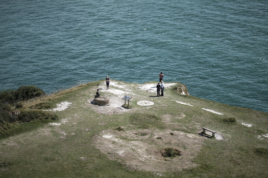Aerial view of people gathered on a grassy coastal peninsula surrounded by turquoise water.