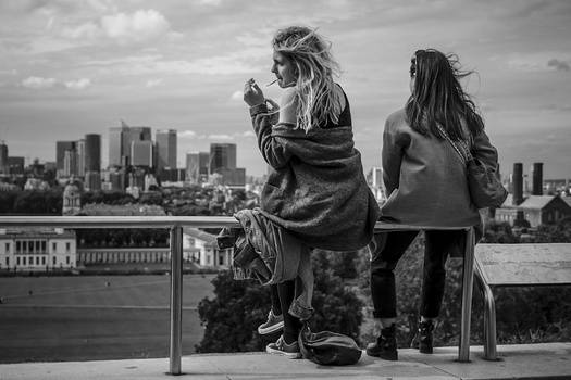 Two people sitting on a railing overlooking a cityscape, one smoking while looking at the view.