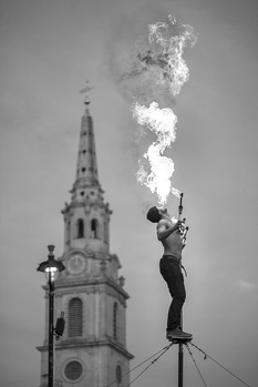 Fire breather performing on a platform with a church tower visible in the background against an overcast sky.