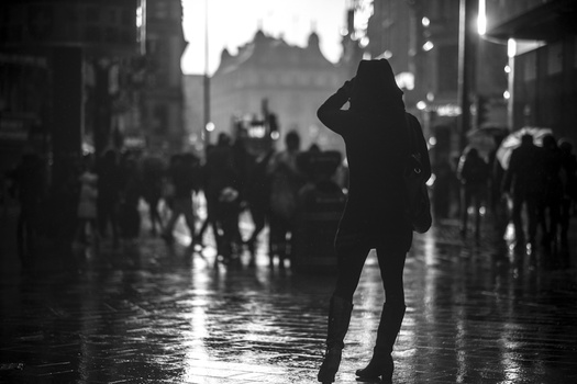 Silhouetted figure standing alone on a wet street amid blurred crowds and city lights.
