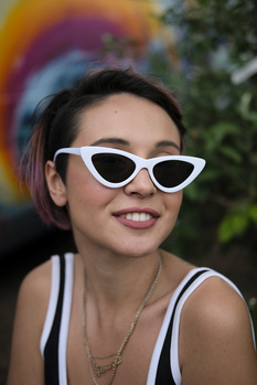 Woman wearing white cat-eye sunglasses and a striped tank top, smiling at camera outdoors.