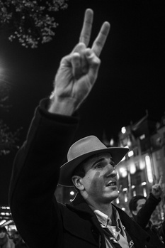 Man in fedora hat making peace sign gesture against dark night sky with lights.
