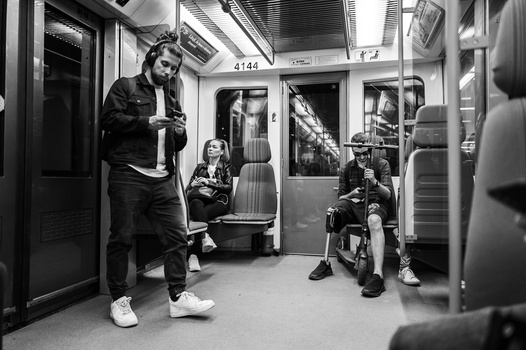Black and white photograph of passengers inside a transit train car, with a standing man in foreground looking at his phone.