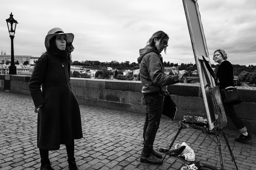 Three people on a stone bridge with a large mirror, one woman in dark coat looking away, another adjusting the mirror, a third on the right.