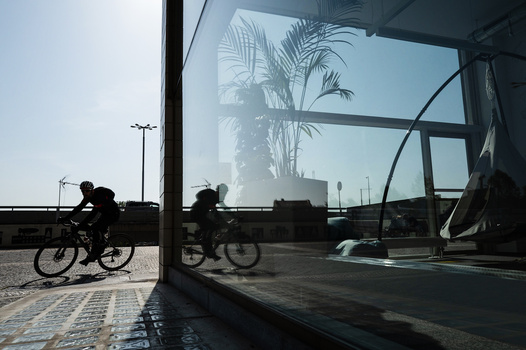 Silhouetted cyclists on a paved promenade next to a modern glass structure reflecting a palm tree against bright sky.