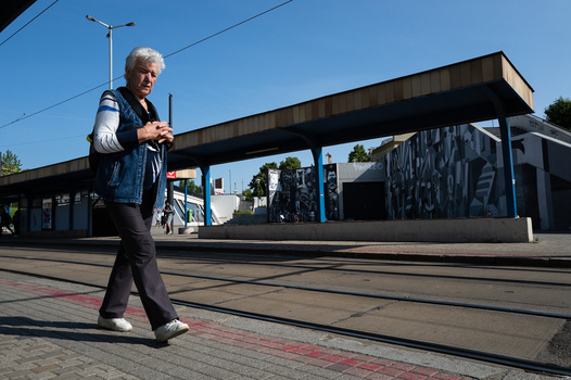 Older woman with white hair walking across tram tracks in an urban transit station on a clear day.