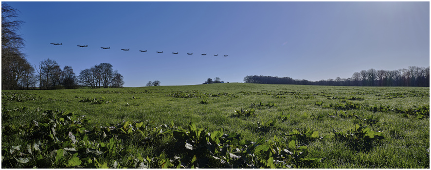 Military aircraft flying in formation over a rural green field under clear blue sky.