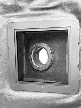 Close-up of a circular lens opening mounted in a square white housing against textured gray fabric.