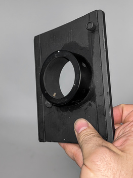 Hand holding a black square filter holder with a circular lens opening against a neutral gray background.