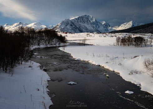 Snow-covered valley with partially frozen river and snow-capped mountains under dramatic sky.