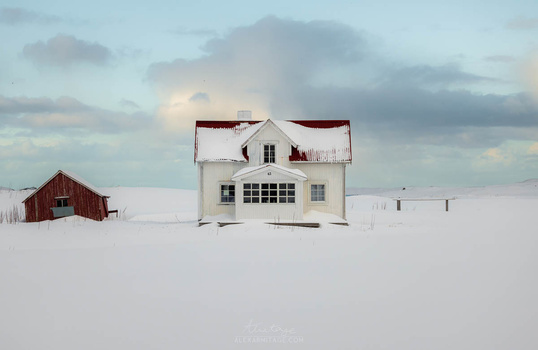 Abandoned cream and red farmhouse standing alone in a vast snowy landscape.