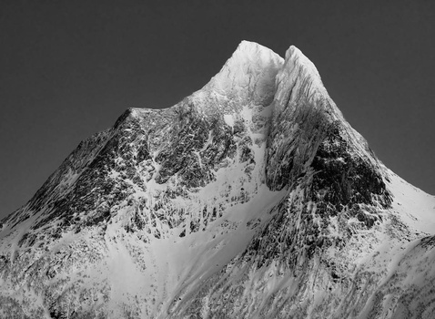 Snow-covered mountain peak with steep rocky ridges against a gray sky.