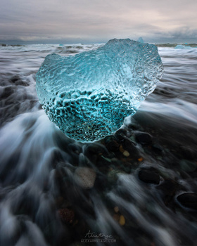 Translucent turquoise ice chunk on black sand beach with waves in motion blur.