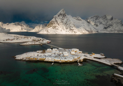 Snow-covered Arctic village on a peninsula surrounded by dark water and jagged mountains under overcast sky.