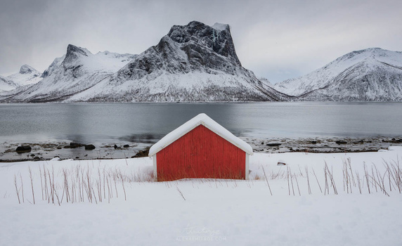 Red house with white roof sitting in snowy field beside frozen lake and snow-capped mountains.