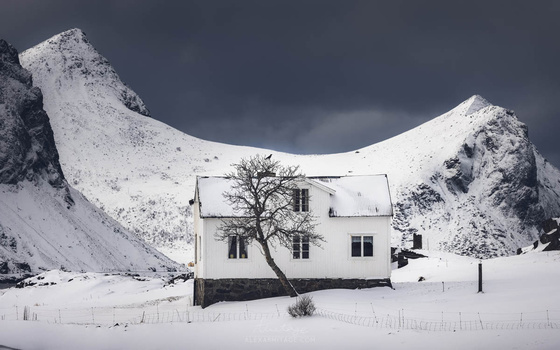 White house with bare tree surrounded by snow-covered mountains under dramatic dark sky.