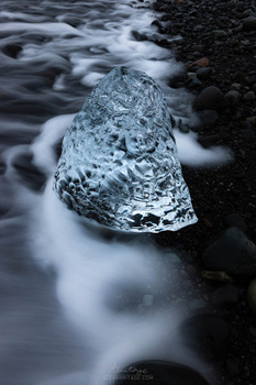Translucent ice chunk resting in flowing river water surrounded by dark rocks.