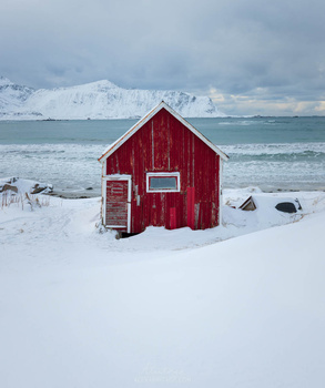 Bright red wooden cabin on a snowy beach with snow-covered mountains across the water in the background.