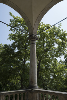 Underside view of a classical stone archway with supporting column and green foliage visible through the opening.
