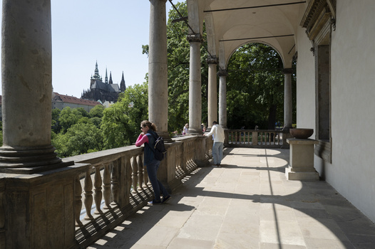 Covered colonnade with classical columns overlooking a distant gothic cathedral tower through green foliage.