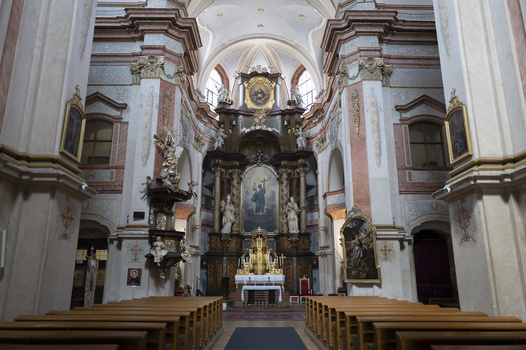 Interior of a baroque church with ornate white and gold altar, symmetrical wooden pews, and vaulted ceiling.