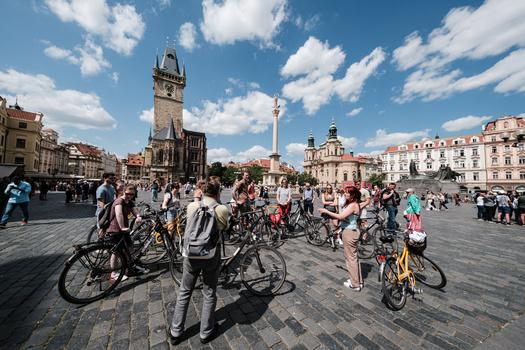 Large group of cyclists gathered in a historic European town square on a sunny day.