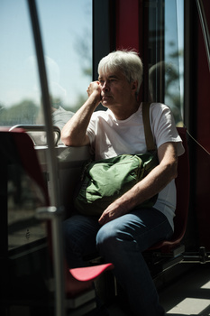 Older man with white hair and glasses sitting by a window on public transportation, gazing thoughtfully outside.