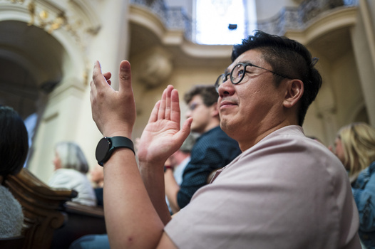 Man with glasses praying with hands raised in an ornate interior space with other worshippers.