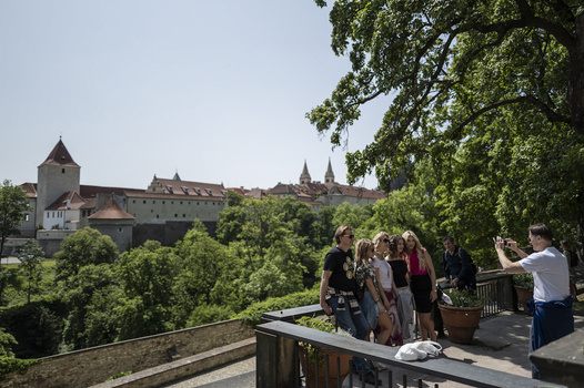 Group of tourists photographing a medieval castle on a hilltop surrounded by dense green forest.