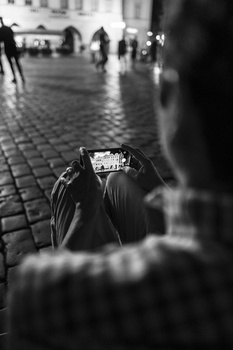 Blurred person in foreground holding smartphone toward camera in urban plaza with pedestrians and wet pavement in soft focus background.