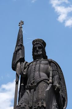 Bronze statue of a bearded man in medieval armor holding a tall staff, photographed against a blue sky.