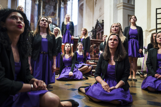 Group of women in purple dresses seated and standing in an ornate interior space, looking upward.