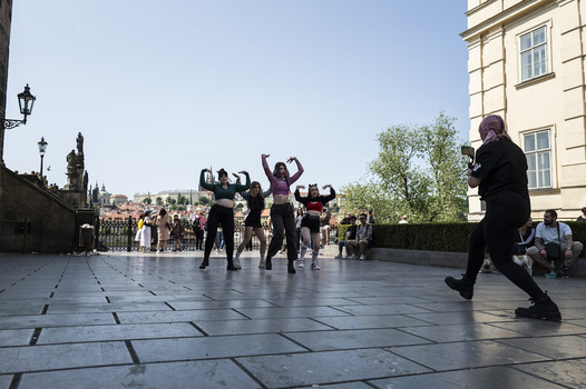 Man jogging across a sunlit plaza while a group of people in dark clothing pose with arms raised in the background.