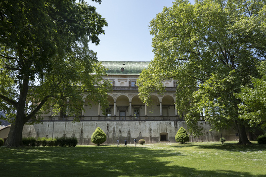Classical stone building with columned portico framed by large trees in manicured grounds.