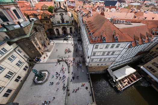 Aerial view of a European town square with terracotta roofs, historic architecture, and a river visible on the right side.