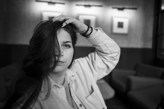 Woman with long dark hair posing indoors, hand raised to her head, wearing a light-colored button-up shirt.