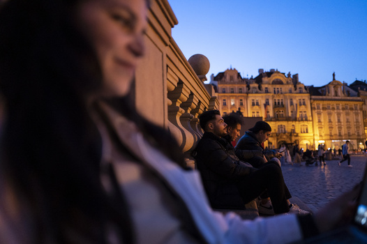People sitting on steps in foreground with European architecture illuminated at dusk in background.