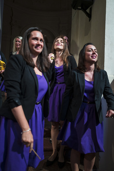 Four women in purple dresses and black blazers smiling at an indoor event with stage lighting.