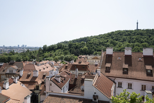 Cityscape with terracotta-roofed buildings in foreground, forested hill with tower in middle distance, and urban skyline under hazy sky.