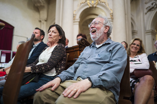 Man in blue shirt smiling while seated in a pew during an indoor gathering at a church or formal venue.
